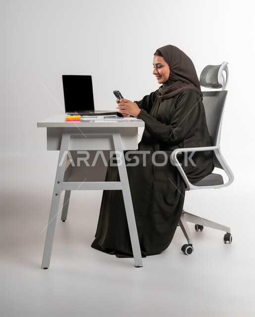 التسوق الالكتروني عبر برامج الهاتف المحمول، مهن وظائف مكتبية نسائية، التواصل والرد على العملاء، دمج التقنية والتكنولوجيا في الحياة العملية، بورتريه لامرأة عربية خليجية سعودية محجبة مبتسمة تجلس امام الطاولة تقوم باستخدام الجوال، خلفية بيضاء
