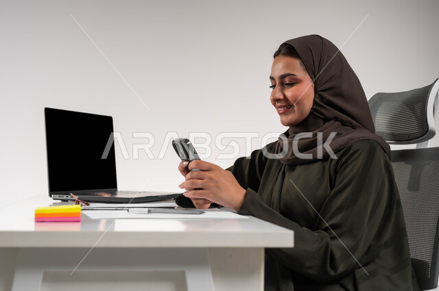 Professions: Women's office jobs, communicating and responding to customers, integrating technology and technology into practical life, online shopping via mobile phone programs, close-up portrait of a veiled Saudi Arabian Gulf woman, smiling, sitting in front of the table using a mobile phone, white background.
