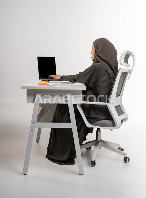 مهن ووظائف نسائية سعوديه، التواصل مع العملاء عبر الجوال، دمج التقنية والتكنولوجيا في الحياة العملية، إجراء مكالمة عمل في مقر الشركة، بورتريه لسيدة أعمال عربية خليجية سعودية محجبة تجلس على الطاولة وتعمل على الحاسوب المحمول، إنجاز المهام المكتبية، خلفية بيضاء