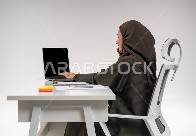 Making a business call at the company headquarters, Saudi women’s professions and jobs, communicating with customers via mobile phone, integrating technology and technology into practical life, close-up portrait of a veiled Saudi Gulf Arab businesswoman sitting at the table and working on the laptop, completing office tasks, white background