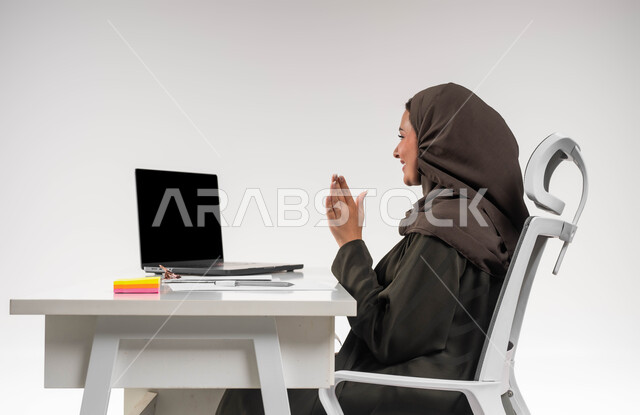 Interaction while attending university lectures online, professions and jobs for Saudi women, managing office work via modern devices remotely, a close-up portrait of a veiled Saudi Arabian Gulf woman wearing an abaya, conducting a live broadcast via the laptop, white background.