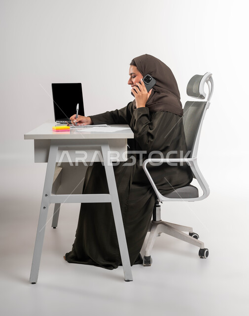 Integrating technology and technology into practical life, making a business call at the company’s headquarters, women’s professions and jobs, communicating with customers via mobile phone, a side portrait of a veiled Saudi Gulf Arab businesswoman sitting at the table recording notes and completing office tasks, white background