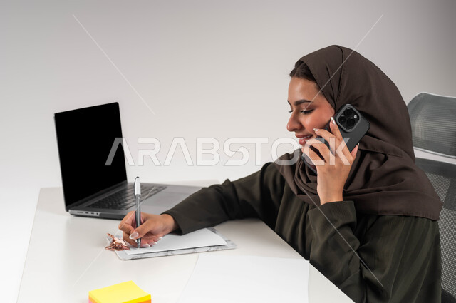 إجراء مكالمة عمل في مقر الشركة، مهن ووظائف نسائية سعوديه، التواصل مع العملاء عبر الجوال، دمج التقنية والتكنولوجيا في الحياة العملية، بورتريه مقرب لسيدة أعمال عربية خليجية سعودية محجبة تجلس على الطاولة تقوم بتسجيل الملاحظات وإنجاز المهام المكتبية، خلفية بيضاء
