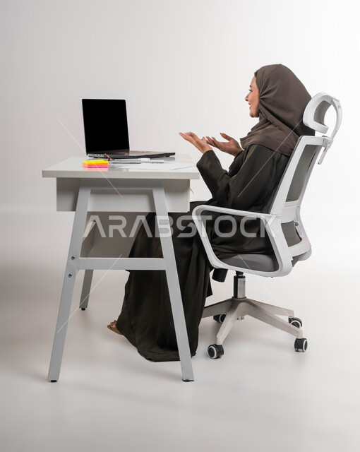 Making video calls and live broadcasting via the laptop, female office job professions, communicating and responding to customers, integrating technology and technology into practical life, a portrait of a veiled Saudi Arabian Gulf woman, smiling, sitting in front of the table using the laptop, white background.