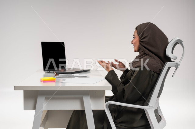 التواصل والرد على العملاء، إجراء مكالمات الفيديو والبث المباشر عبر اللابتوب، مهن وظائف مكتبية نسائية، دمج التقنية والتكنولوجيا في الحياة العملية، بورتريه مقرب من الجانب لامرأة عربية خليجية سعودية محجبة تجلس امام الطاولة تقوم باستخدام الحاسوب المحمول، خلفية بيضاء