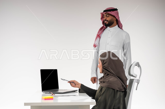 دمج العمل مع التقنية والتكنولوجيا، الاحترافية في أداء المهام للتطور المهني و تحقيق الأهداف، بورتريه مقرب لزميلين عربيين خليجيين سعوديين مبتسمين يقومان بإنجاز المهام عبر الحاسوب المحمول، الاشارة الى اللابتوب، العمل الجماعي و روح الفريق الواحد، خلفيه بيضاء