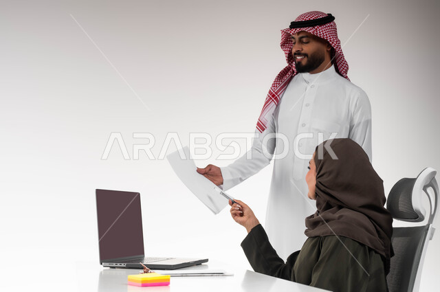 Delivering tasks and projects on time, integrating work with technology and technology, a close-up portrait of two smiling Gulf Arab Saudi colleagues exchanging conversations about successful deals, cooperation and teamwork, directing efforts to achieve the company’s goals, white background.