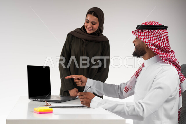 تبادل الأفكار والخبرات لتحقيق الأهداف، بورتريه لزميلين عربيين خليجيين سعوديين مبتسمين يقومان بإنجاز المهام عبر الحاسوب المحمول داخل المكتب، الاشارة الى اللابتوب والتنبيه لتطبيق الخطط، التعاون وروح الفريق الواحد، تعليم الموظفين أساليب العمل الجديدة، خلفيه بيضاء