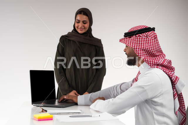تدريب الموظفين على الأساليب الجديدة في العمل لتحقيق أهداف الشركة، دمج البيئة العملية مع التقنية والتكنولوجيا، بورتريه مقرب لزميلين عربيين خليجيين سعوديين مبتسمين يقومان بإنجاز المهام عبر الحاسوب المحمول داخل المكتب، التعاون وروح الفريق الواحد، خلفيه بيضاء
