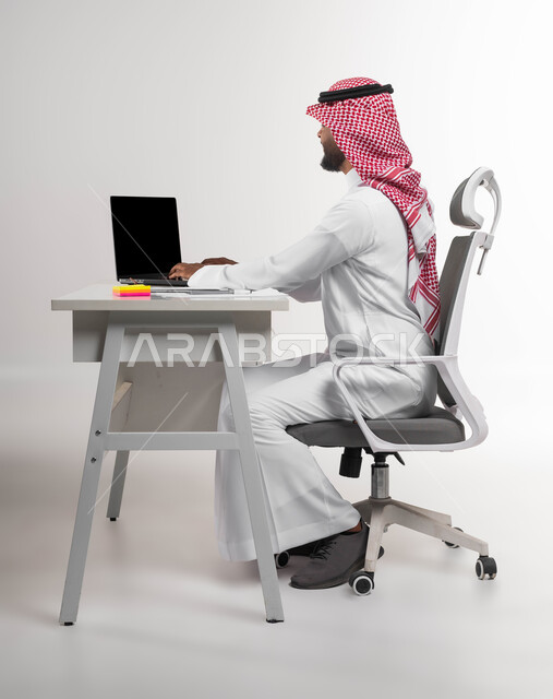 مهن ووظائف مكتبية سعودية، مفهوم ادارة الاعمال وتحقيق اهداف الشركة، بورتريه من الجانب لرجل اعمال عربي خليجي سعودي يرتدي الشماغ والثوب التقليدي يجلس في مكتبه يعمل على الحاسوب المحمول بايماءات الحماس والسعادة، انجاز مهام العمل عن بعد، خلفية بيضاء
