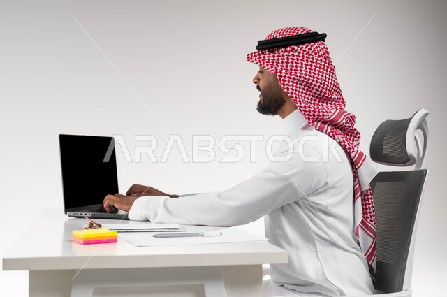 مفهوم ادارة الاعمال وتحقيق اهداف الشركة، بورتريه مقرب من الجانب لرجل اعمال عربي خليجي سعودي يرتدي الشماغ والثوب التقليدي يجلس في مكتبه يعمل على الحاسوب المحمول بايماءات الحماس والسعادة، انجاز مهام العمل عن بعد، مهن ووظائف مكتبية سعودية، خلفية بيضاء