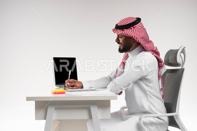 كتابة الملاحظات وتطوير الخطط لتحقيق اهداف الشركة، بورتريه من الجانب لرجل عربي خليجي سعودي مبتسم يرتدي الشماغ والثوب التقليدي يجلس في مكتبه يعمل على الحاسوب المحمول، تدوين وتسجيل بيانات ومعلومات العمل، مفهوم ادارة الاعمال، مهن ووظائف مكتبية سعودية، خلفية بيضاء