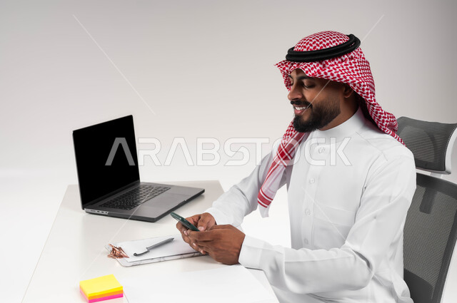 التواصل مع الاصدقاء في وقت العمل، بورتريه مقرب لرجل عربي خليجي سعودي مبتسم يرتدي الشماغ والثوب التقليدي يجلس في مكتبه ويقوم بإستخدام الهاتف المحمول ومتابعة أخبار الشركات العالمية، مهن ووظائف مكتبية سعودية، تصفح وسائل التواصل الاجتماعي، خلفية بيضاء