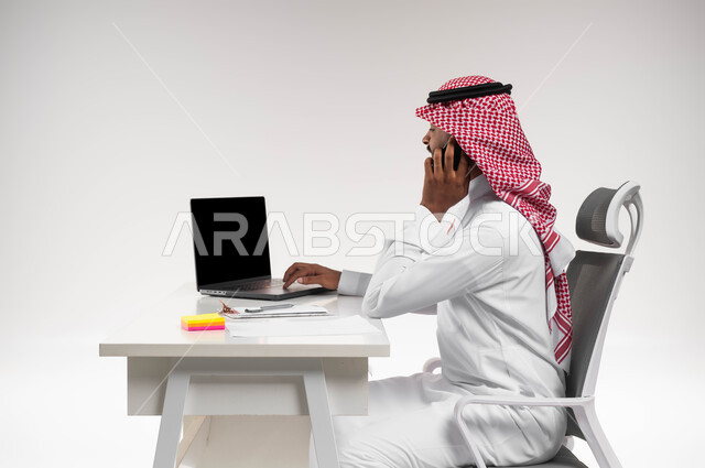 الرد والاجابة على الاستفسارات والاسئلة، بورتريه مقرب من الجانب لرجل عربي خليجي سعودي يرتدي الشماغ والثوب التقليدي يجلس في مكتبه ويقوم بإجراء مكالمة هاتفية، مفهوم خدمة الزبائن والعملاء، إدخال المعلومات والبيانات الخاصة بالشركة على الحاسوب، خلفية بيضاء