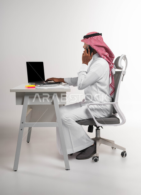 مفهوم خدمة الزبائن والعملاء، الرد والاجابة على الاستفسارات والاسئلة، بورتريه من الجانب لرجل عربي خليجي سعودي يرتدي الشماغ والثوب التقليدي يجلس في مكتبه ويقوم بإجراء مكالمة هاتفية، إدخال المعلومات والبيانات الخاصة بالشركة على الحاسوب، خلفية بيضاء