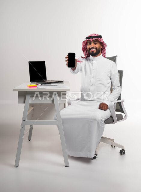Completing tasks online, displaying company websites and services via mobile phone, portrait of a smiling Saudi Arabian Gulf man wearing a shemagh and traditional thobe, sitting in his office, displaying a blank black screen on the mobile phone looking at the camera, integrating work with technology and technology, white background