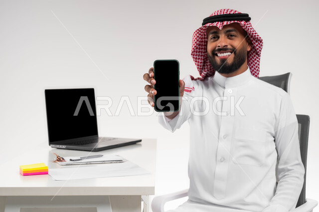 عرض مواقع وخدمات الشركة عبر الجوال, انجاز المهام عبر الانترنت, بورتريه مقرب لرجل عربي خليجي سعودي مبتسم يرتدي الشماغ والثوب التقليدي يجلس في مكتبه ويقوم بعرض شاشة سوداء فارغة عبر الهاتف المحمول ينظر الى الكاميرا, دمج العمل مع التقنية والتكنولوجيا، خلفية بيضاء