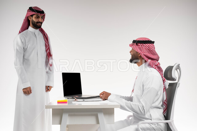 The desire to learn work methods and professional development, a close-up portrait of two Saudi Gulf Arab employees completing tasks and work using a laptop computer in the office, the concept of teamwork and one team spirit, developing plans and solving problems to achieve the company’s goals, white background.