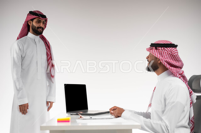 Developing plans and solving problems to achieve the company’s goals, the desire to learn work methods and professional development, a close-up portrait of two Saudi Gulf Arab employees completing tasks and work using the laptop in the office, the concept of teamwork and the spirit of one team, a white background.