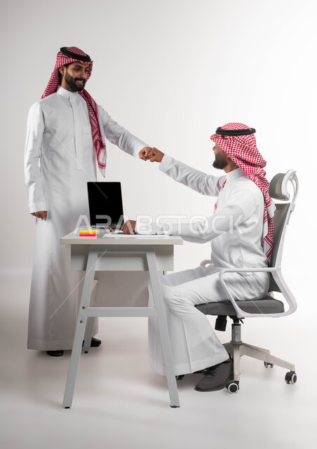 Getting acquainted and building social relationships, exchanging experiences and successful agreements, a portrait of smiling Gulf Arab Saudi employees wearing traditional Saudi dress greeting each other by holding hands inside the office, expressions of deep friendship, greeting without contact to prevent the Corona virus, white background.