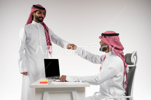 تعابير الصداقة العميقة، تبادل الخبرات والاتفاقيات الناجحة في مكتب الشركه، مفهوم الإدارة المتعاونة، بورتريه مقرب لصديقين عربيين خليجيين سعوديين مبتسمين يرتديان الشماغ و الثوب التقليدي يقومان بالسلام بقبضة اليد، خلفية بيضاء.