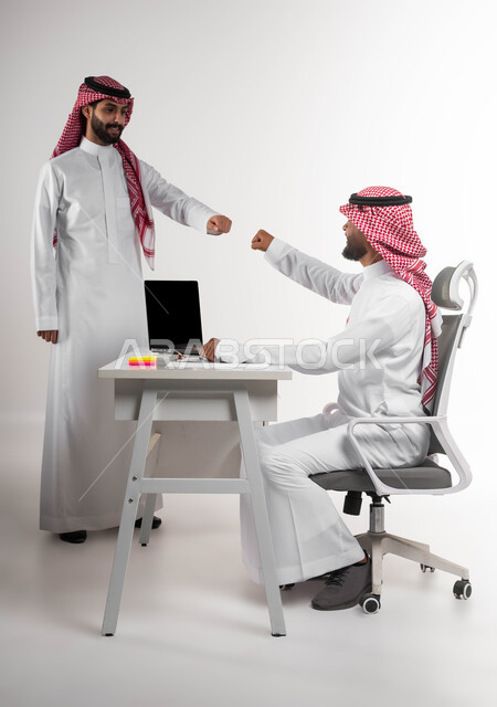 التحية في وباء الكورونا، تعابير الصداقة العميقة، تبادل الخبرات والاتفاقيات الناجحة في مكتب الشركه، مفهوم الإدارة المتعاونة، بورتريه مقرب لصديقين عربيين خليجيين سعوديين مبتسمين يرتديان الشماغ والثوب التقليدي يقومان بالسلام بقبضة اليد بدون ملامسة، خلفية بيضاء.