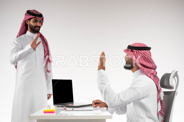 تبادل الخبرات والاتفاقيات الناجحة في مكتب الشركه، مفهوم الإدارة المتعاونة بين الموظفين، بورتريه مقرب لصديقين عربيين خليجيين سعوديين مبتسمين يرتديان الشماغ والثوب التقليدي يقومان بالتلويح باليد والسلام عن بعد، تعابير الصداقة العميقة في العمل، خلفية بيضاء.