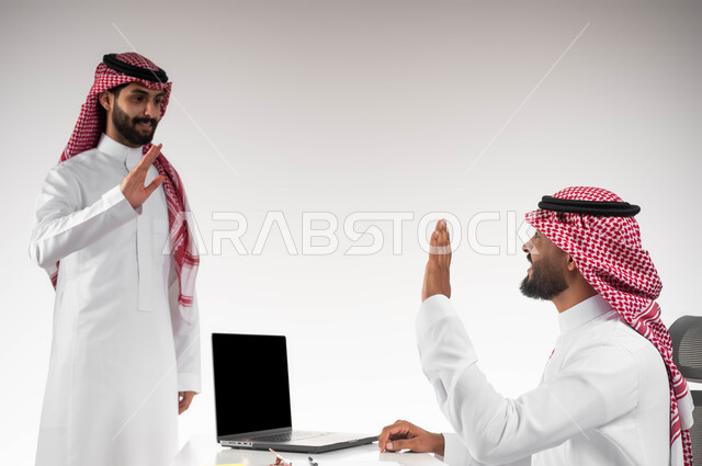 تبادل الخبرات والاتفاقيات الناجحة في مكتب الشركه، مفهوم الإدارة المتعاونة بين الموظفين، بورتريه مقرب لصديقين عربيين خليجيين سعوديين مبتسمين يرتديان الشماغ والثوب التقليدي يقومان بالتلويح باليد والسلام عن بعد، خلفية بيضاء.