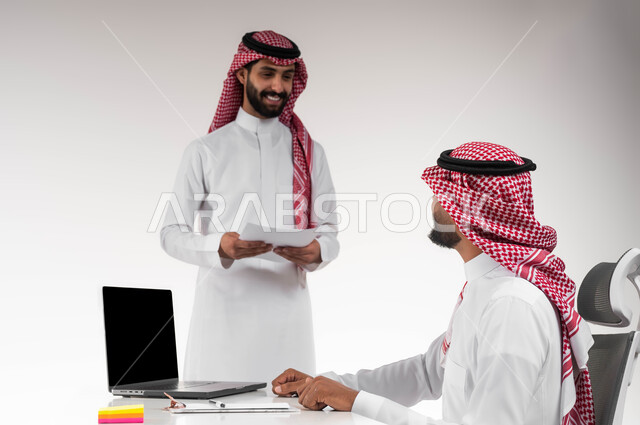 عرض صفقة عمل جديدة، إنجاز المهام اليومية بالوقت اللازم، بورتريه لموظفين عربيين خليجيين سعوديين مبتسمين يرتديان الشماغ والثوب التقليدي يتناقشان في أمور العمل باستخدام الأوراق والبيانات، الوحدة والتفاعل الجماعي لتحقيق أهداف الشركة وحل المشكلات، خلفية بيضاء.