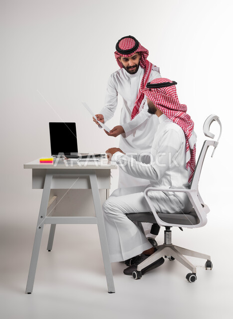 Explaining new work methods to employees, discussing work matters using papers and data, a portrait of two smiling Saudi Gulf Arab men wearing the traditional shemagh and thobe, concluding a new partnership deal, unity and group interaction to achieve the company’s goals and solve problems, white background.