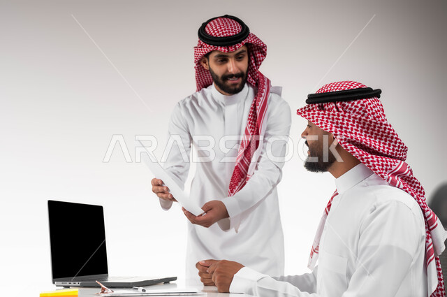 الرغبة في تعلم أساليب العمل و التطور المهني، بورتريه مقرب لموظفين عربيين خليجيين سعوديين مبتسمين يرتديان الشماغ والثوب التقليدي يقومان بمراجعة البيانات من خلال الورق، تحقيق أهداف الشركة وحل المشكلات، عرض مزايا الصفقات الجديدة، خلفية بيضاء.