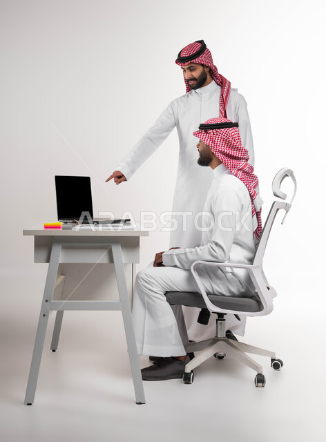 مفهوم الإدارة المتعاونة، الإشارة إلى شاشة الحاسوب، التفاعل الجماعي والوحدة لتحقيق أهداف الشركة وحل المشكلات، بورتريه من الجانب لموظفين عربيين خليجيين سعوديين مبتسمين يرتديان الشماغ والثوب التقليدي يناقشان أمور العمل، خلفية بيضاء.