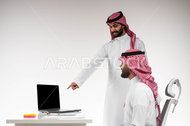 الإشارة إلى شاشة الحاسوب، التفاعل الجماعي والوحدة لتحقيق أهداف الشركة وحل المشكلات، مفهوم الإدارة المتعاونة، بورتريه مقرب من الجانب لموظفين عربيين خليجيين سعوديين مبتسمين يرتديان الشماغ والثوب التقليدي يناقشان أمور العمل، خلفية بيضاء.