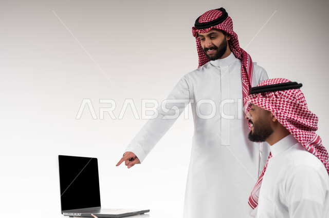Group interaction and unity to achieve the company's goals and solve problems, pointing to the computer screen, the concept of cooperative management, a close-up portrait from the side of two smiling Saudi Gulf Arab employees wearing the shemagh and traditional thobe discussing work matters, white background