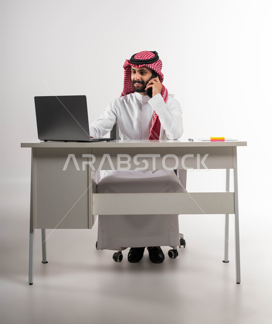 إنجاز المهام المكتبية، مكالمة عمل في مقر الشركة، بورتريه لشاب عربي سعودي خليجي مبتسم يرتدي الشماغ والثوب التقليدي يمسك الهاتف المحمول ويقوم بإجراء مكالمة هاتفيه, استخدام جهاز اللابتوب والتقنيات الحديثة، خلفية بيضاء .