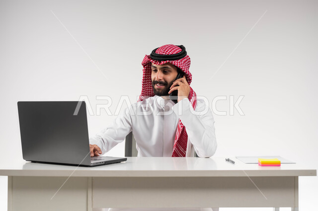 استخدام جهاز اللابتوب والتقنيات الحديثة، مكالمة عمل في مقر الشركة، بورتريه مقرب لشاب عربي سعودي خليجي مبتسم يرتدي الشماغ والثوب التقليدي يمسك الهاتف المحمول ويقوم بإجراء مكالمة هاتفيه, إنجاز المهام المكتبية، خلفية بيضاء .