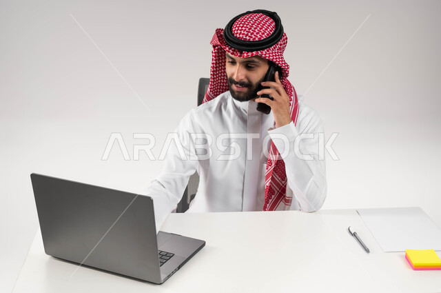 مكالمة عمل في مقر الشركة، استخدام جهاز اللابتوب والتقنيات الحديثة، بورتريه مقرب لشاب عربي سعودي خليجي مبتسم يرتدي الشماغ والثوب التقليدي يمسك الهاتف المحمول ويقوم بإجراء مكالمة هاتفيه, إنجاز المهام المكتبية، خلفية بيضاء .