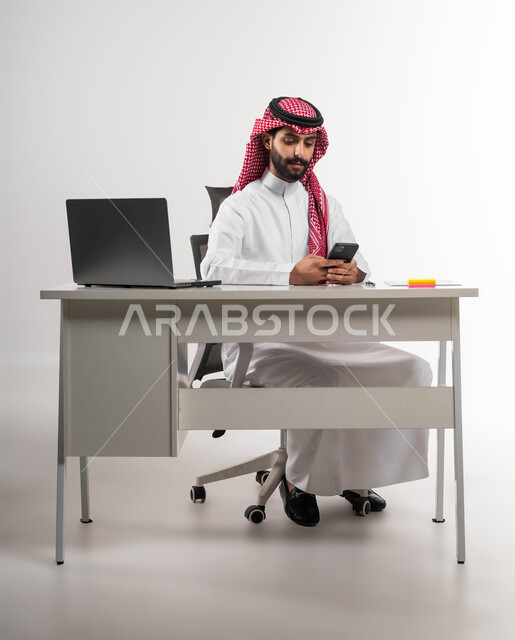 Browsing social networking sites, using modern technologies, online shopping via mobile phone programs, a portrait of a smiling Saudi Arabian Gulf man wearing a shemagh and traditional dress, holding a mobile phone and chatting with family and friends, white background.