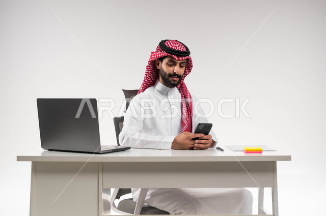 استخدام التقنيات الحديثة، التسوق الالكتروني عبر برامج الهاتف المحمول، الدردشة مع الأهل والأصدقاء، بورتريه لرجل عربي سعودي خليجي مبتسم يرتدي الشماغ والثوب التقليدي يمسك الجوال ويتصفح مواقع التواصل الاجتماعي, خلفية بيضاء .
