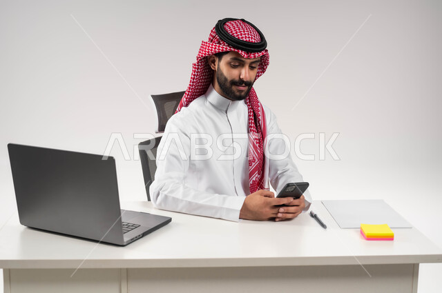 Online shopping via mobile phone programs, chatting with family and friends, a close-up portrait of a smiling Saudi Arabian Gulf man wearing a shemagh and traditional dress, holding a mobile phone and browsing social media sites, using modern technologies, a white background