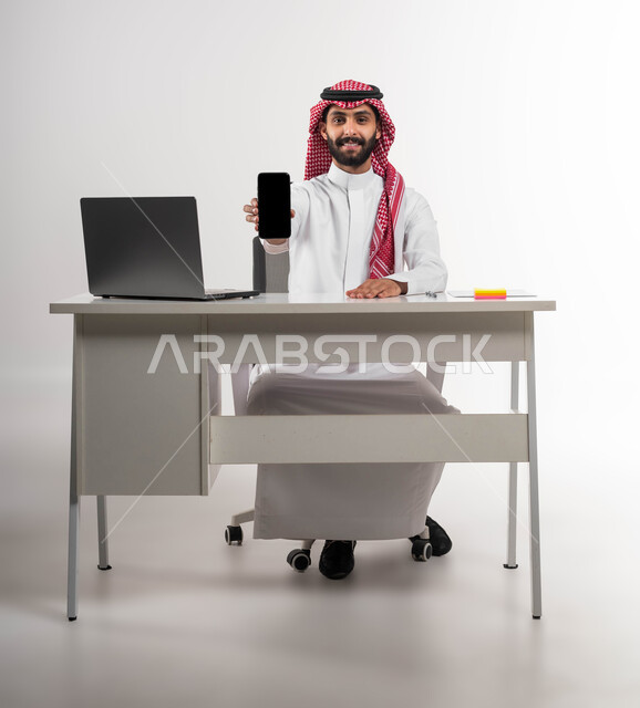 الاهتمام بالأناقة والمظهر الخارجي، عرض تطبيقات وبرامج الهاتف المحمول، إيماءات الرجولة والثقة بالنفس، بورتريه لرجل عربي سعودي خليجي مبتسم يرتدي الشماغ والثوب التقليدي ينظر إلى الكاميرا ويمسك الجوال بشاشة سوداء فارغة, خلفية بيضاء .