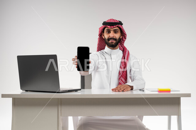 عرض تطبيقات وبرامج الهاتف المحمول، إيماءات الرجولة والثقة بالنفس، بورتريه لرجل عربي سعودي خليجي مبتسم يرتدي الشماغ والثوب التقليدي ينظر إلى الكاميرا يمسك الجوال بشاشة سوداء فارغة, الاهتمام بالأناقة والمظهر الخارجي، خلفية بيضاء .
