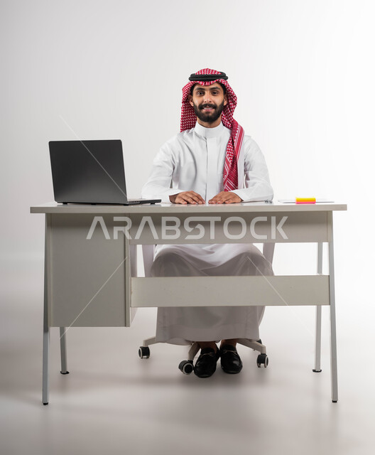 إيماءات الرجولة والثقة بالنفس، شخصية بارزة ومهمة، بورتريه  لشاب عربي سعودي خليجي مبتسم يرتدي الشماغ والثوب التقليدي يجلس على كرسي المكتب ينظر إلى الكاميرا, الاهتمام بالأناقة والمظهر الخارجي، خلفية بيضاء .