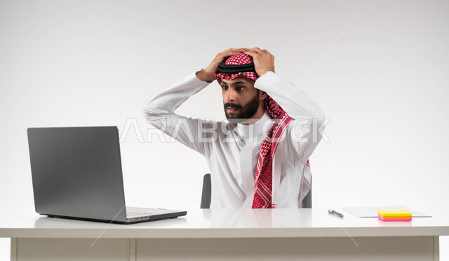 خسارة الشركة والفشل في تحقيق الأهداف, الشعور بالانزعاج والصدمة, تعابير الاندهاش بسماع الاخبار السيئة, بورتريه  مقرب لرجل عربي سعودي خليجي يرتدي الشماغ والثوب التقليدي يضع يديه على رأسه بإيماءات الذهول يقوم باستخدام جهاز الحاسوب المحمول, خلفية بيضاء .