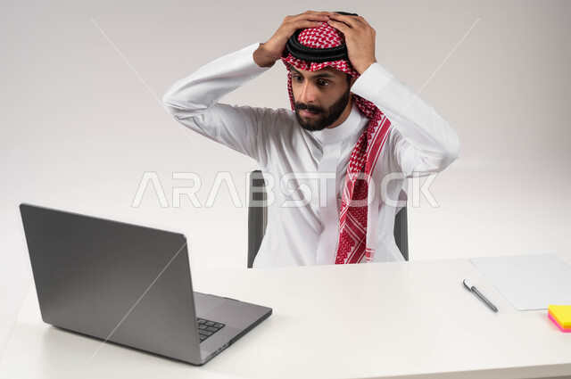 الخسارة والفشل في تحقيق الأهداف, الشعور بالانزعاج والصدمة, تعابير الاندهاش بسماع الاخبار السيئة, بورتريه  مقرب لرجل عربي سعودي خليجي يرتدي الشماغ والثوب التقليدي يضع يديه على رأسه بإيماءات الذهول يقوم باستخدام جهاز الحاسوب المحمول, خلفية بيضاء .