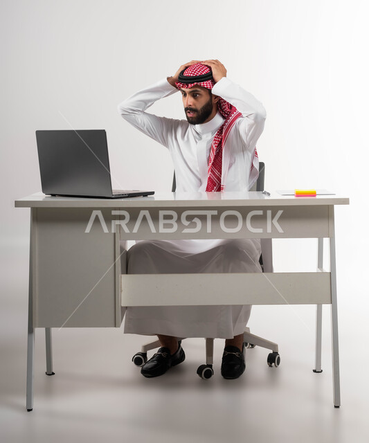 الشعور بالانزعاج والصدمة, تعابير الاندهاش بسماع الاخبار السيئة, الفشل في تحقيق الأهداف, بورتريه لرجل عربي سعودي خليجي يرتدي الشماغ والثوب التقليدي يضع يديه على رأسه بإيماءات الذهول يقوم باستخدام جهاز الحاسوب المحمول, خلفية بيضاء .