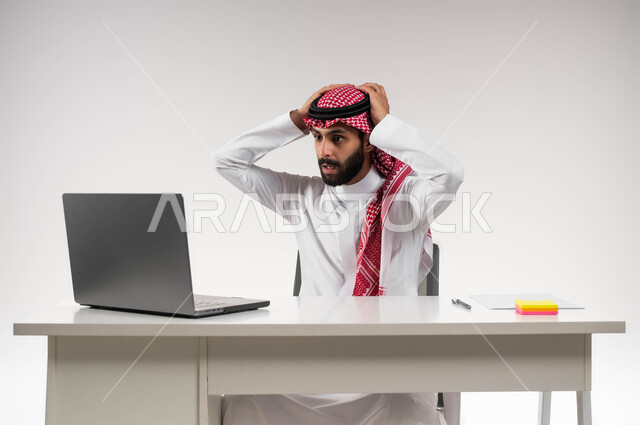 الفشل في تحقيق الأهداف, تعابير الاندهاش بسماع الاخبار السيئة, بورتريه لرجل عربي سعودي خليجي يرتدي الشماغ والثوب التقليدي يضع يديه على رأسه بإيماءات الذهول يقوم باستخدام جهاز الحاسوب المحمول, الشعور بالانزعاج والصدمة, خلفية بيضاء .