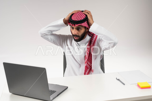 تعابير الاندهاش بسماع الاخبار السيئة, الفشل في تحقيق الأهداف, بورتريه  مقرب لرجل عربي سعودي خليجي يرتدي الشماغ والثوب التقليدي يضع يديه على رأسه بإيماءات الذهول يقوم باستخدام جهاز الحاسوب المحمول, الشعور بالانزعاج والصدمة, خلفية بيضاء .