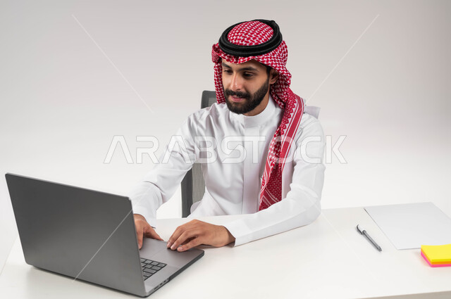 النقر والتفاعل مع لوحة المفاتيح, التفكير في استراتيجيات وأهداف العمل، بورتريه مقرب لموظف عربي سعودي خليجي يرتدي الشماغ والثوب التقليدي يقوم بالعمل على جهاز اللابتوب المحمول, إيماءات التركيز والتمعن, إدارة شؤون الأعمال وتنظيمها, خلفية بيضاء .