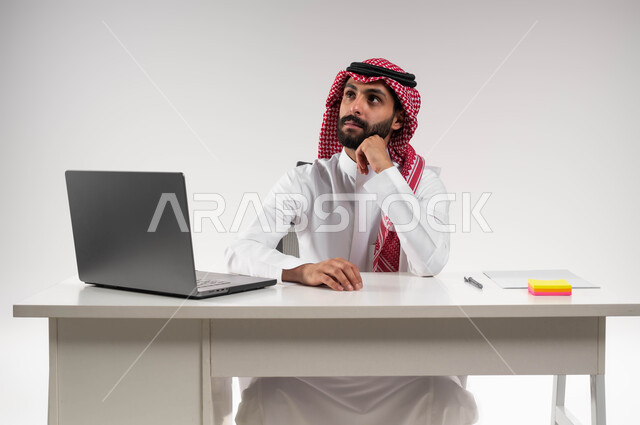 إيماءات التركيز والتمعن, النظر إلى شيء ما في الأعلى, تعابير التفكير في استراتيجيات وأهداف العمل، بورتريه لرجل عربي سعودي خليجي يرتدي الشماغ والثوب التقليدي يقوم بالتخطيط والتأمل داخل مكتبه, إدارة شؤون الأعمال وتنظيمها, خلفية بيضاء .